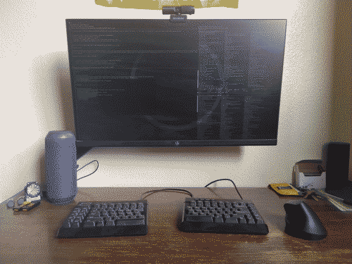Wooden desktop with a split mechanical keyboard, vertical wireless mouse, 27" flat panel on a clamp arm, a sound can, and a small clock and USB drives