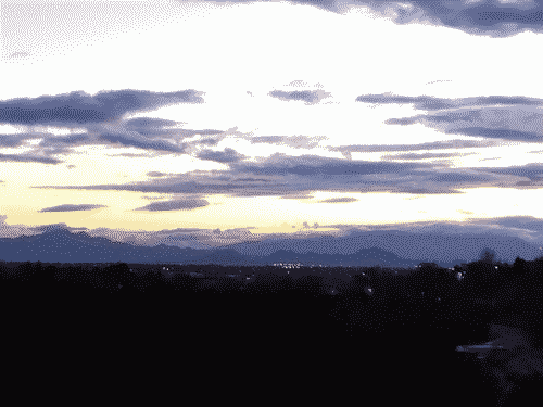 Santa Fe skyline from the north looking southeast at twilight; treeline is in the foreground, the city with its lights in midground, and the background showing brilliant indigo shades of the mountains fading into the low clouds at sunset, with the sky above the mountains and lowest clouds fading from golden into light grey; a smattering of indigo clouds streak across the sky in the upper half of the image