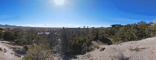 Panoramic shot of the hill on the northside of Santa Fe, scrublands and white sands against blue sky, with the blazing white sun peeking out of the top frame of the shot