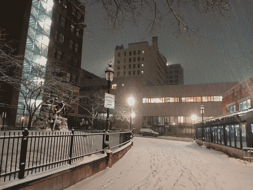 View of downtown Portland, Maine at street level: snow covering the sidewalk leafless trees, multi-level buildings with various levels of lights visible through the windows - on the left side a stairwell is brilliantly bleeding white light into the street; on the right a cafe/bar glows purple, and in front two buildings stagger over the street with yellow and black windows