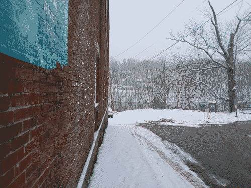 Looking toward the riverside from the 14 State St building, its brick wall on the left of the shot, parking lot descending toward the riverbank with leafless deciduous trees and much snow on the ground