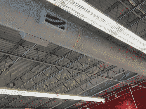 Eggshell corrugated metal cieling with steel tresses running directly under it, and perpendicular to them a large round eggshell HVAC duct between two long flourescent light fixtures that hang on wires. In the bottom-right of the frame the cieling meets the red wall.